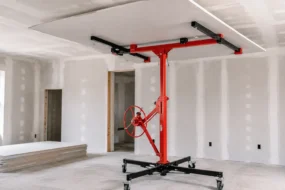 Blog featured image guide on choosing a drywall lift at Harbor Freight, showing a red mechanical panel hoist holding a large sheet of drywall flat against a ceiling during room installation.