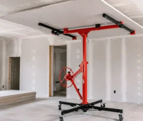 Blog featured image guide on choosing a drywall lift at Harbor Freight, showing a red mechanical panel hoist holding a large sheet of drywall flat against a ceiling during room installation.