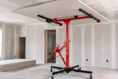 Blog featured image guide on choosing a drywall lift at Harbor Freight, showing a red mechanical panel hoist holding a large sheet of drywall flat against a ceiling during room installation.