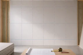 Blog featured image for a drywall layout guide, showing a gridded schematic blue chalk line grid over a large hung drywall room wall, with a blueprint scroll, tape measure, pencil, and stacks of plasterboard panels positioned in the foreground foreground.