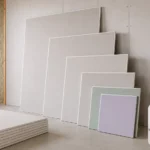 Blog featured image guide on drywall sizes and thickness options, showcasing a stack and staggered lineup of various plasterboard sheets next to an open wall frame, with circular edge-profile insets detailing one-quarter inch, three-eighths inch, one-half inch, and five-eighths inch options alongside a small 3D residential layout diagram.