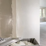 Blog featured image guide on how long does drywall mud take to dry, showcasing a freshly mudded corner wall seam with a clear contrast between wet dark grey joint compound and dry lighter patches, alongside a mud pan and taping knife in the foreground.