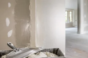 Blog featured image guide on how long does drywall mud take to dry, showcasing a freshly mudded corner wall seam with a clear contrast between wet dark grey joint compound and dry lighter patches, alongside a mud pan and taping knife in the foreground.