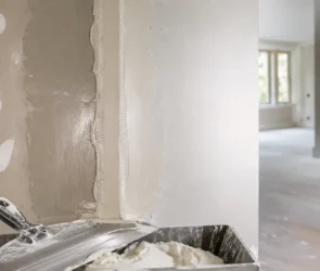 Blog featured image guide on how long does drywall mud take to dry, showcasing a freshly mudded corner wall seam with a clear contrast between wet dark grey joint compound and dry lighter patches, alongside a mud pan and taping knife in the foreground.